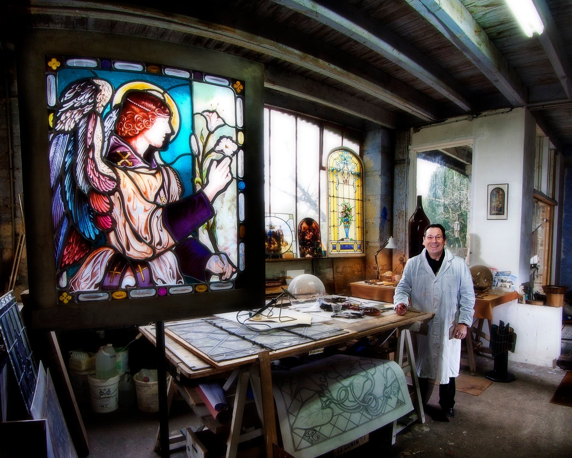 Philippe Audoux, maitre verrier posant dans son atelier, entouré de ses réalisations.