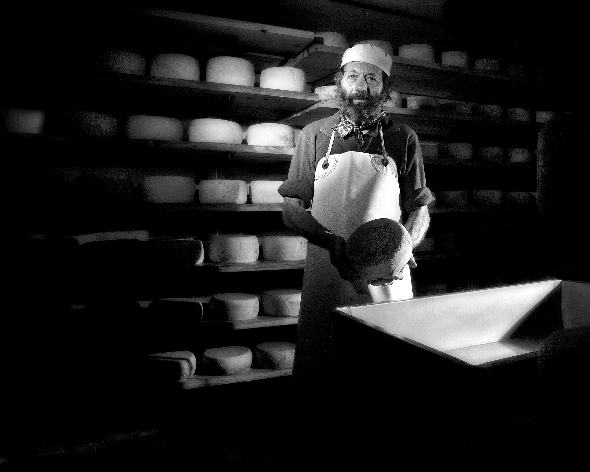 Portrait en noir et blanc d'un fromager tenant une meule devant ses étagères d'affinage